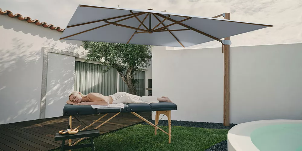 Frau entspannt auf einer Liege unter einem großen, weißen Glatz Aura Sonnenschirm in einem modernen Garten mit Holzterrasse.