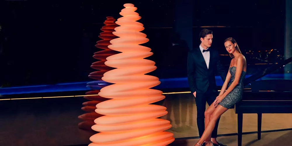 Zwei Personen in Abendkleidung stehen neben einer leuchtenden, spiralförmigen Vondom Chrismy Gartenleuchte in warmem Orange vor dunklem Hintergrund.