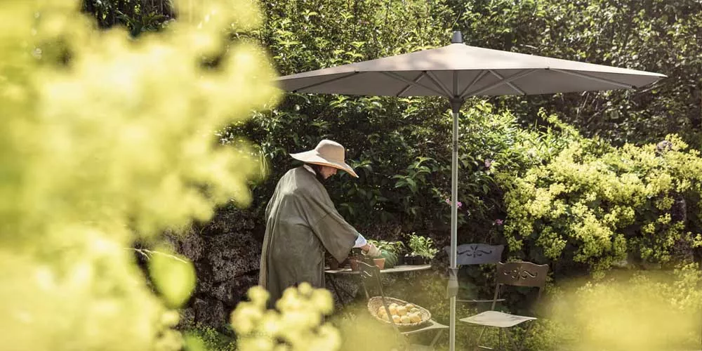 Glatz Alu-Twist Sonnenschirm in Taupe, aufgestellt in einem sonnigen Garten mit blühenden Sträuchern im Hintergrund.