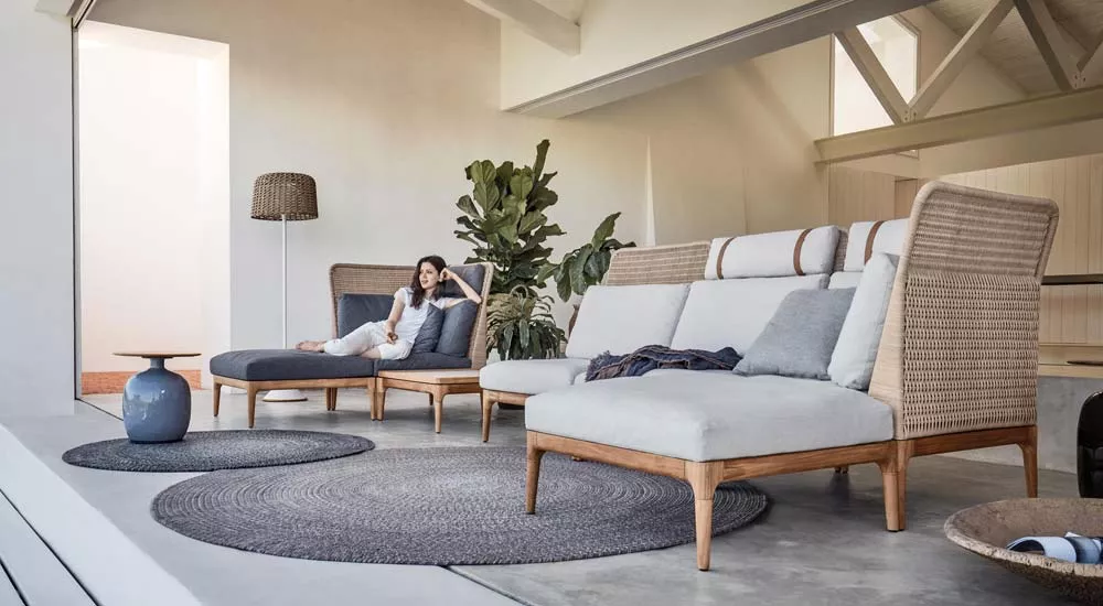 Gloster Lima Gartenmöbel Loungegruppe in Naturtönen auf einer Terrasse mit Pflanzen im Hintergrund, gemütliche Atmosphäre.