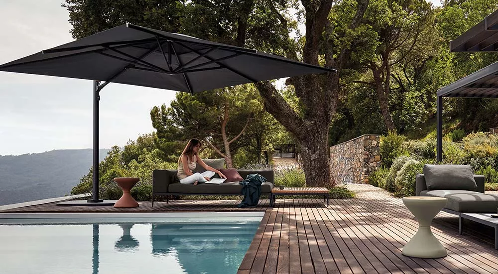 Frau entspannt auf der Gloster Grid Gartenmöbel Loungegruppe in natürlicher Umgebung neben einem Pool unter einem großen Sonnenschirm.