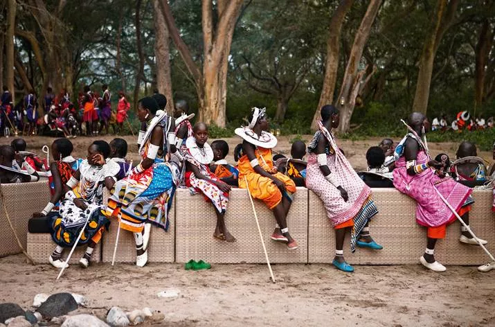 Eine Gruppe von Menschen in traditioneller Kleidung sitzt und steht auf einer Lounge im Freien umgeben von Bäumen und Natur.