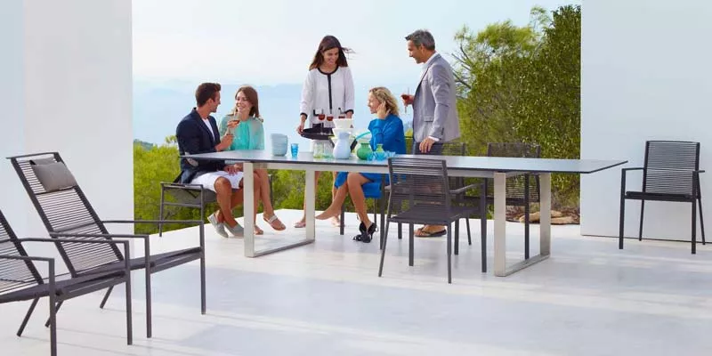 Vier Personen sitzen und stehen um einen Cane-line Edge Tisch in einem modernen, offenen Außenbereich mit minimalistischen Stühlen.