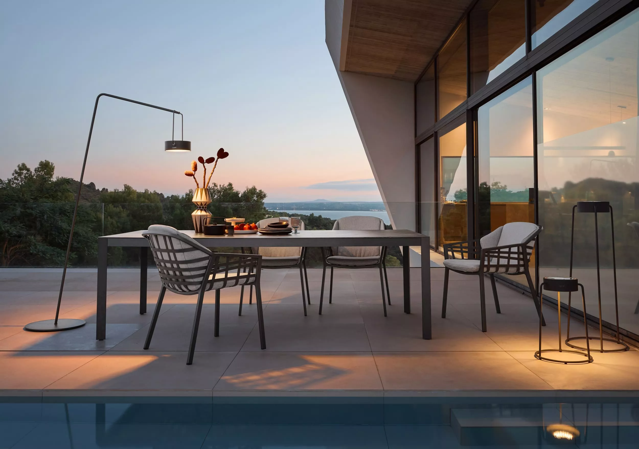 Solpuri Classic Alu Tisch und Stühle auf einer Terrasse bei Sonnenuntergang mit gedecktem Tisch und dekorativer Beleuchtung im Hintergrund.