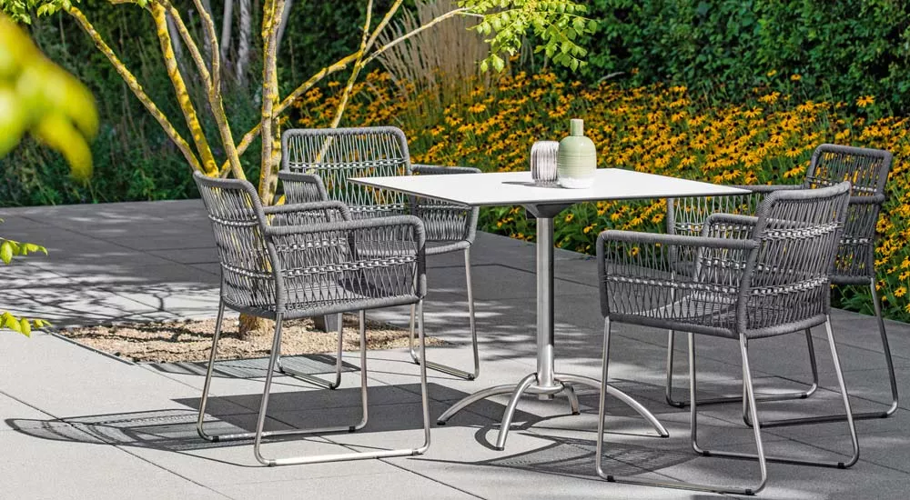 Fischer Möbel Filo Essgruppe in Grau auf einer Terrasse mit blühenden gelben Blumen im Hintergrund.