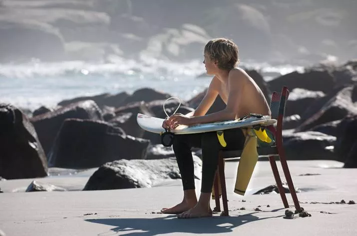 Ein junger Mann sitzt auf einem Dedon Play Essstuhl am Strand und hält ein Surfbrett während er auf das Meer schaut.