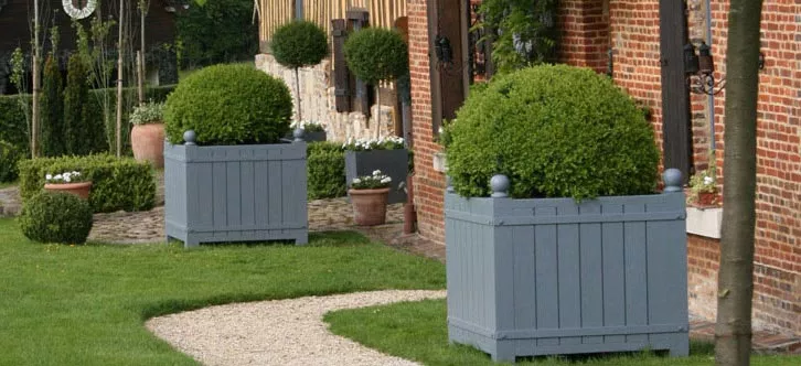 Zwei Orangerie Pflanzkübel aus lackiertem Akazienholz stehen im Garten vor einem Backsteingebäude mit gepflegtem Rasen und Kiesweg