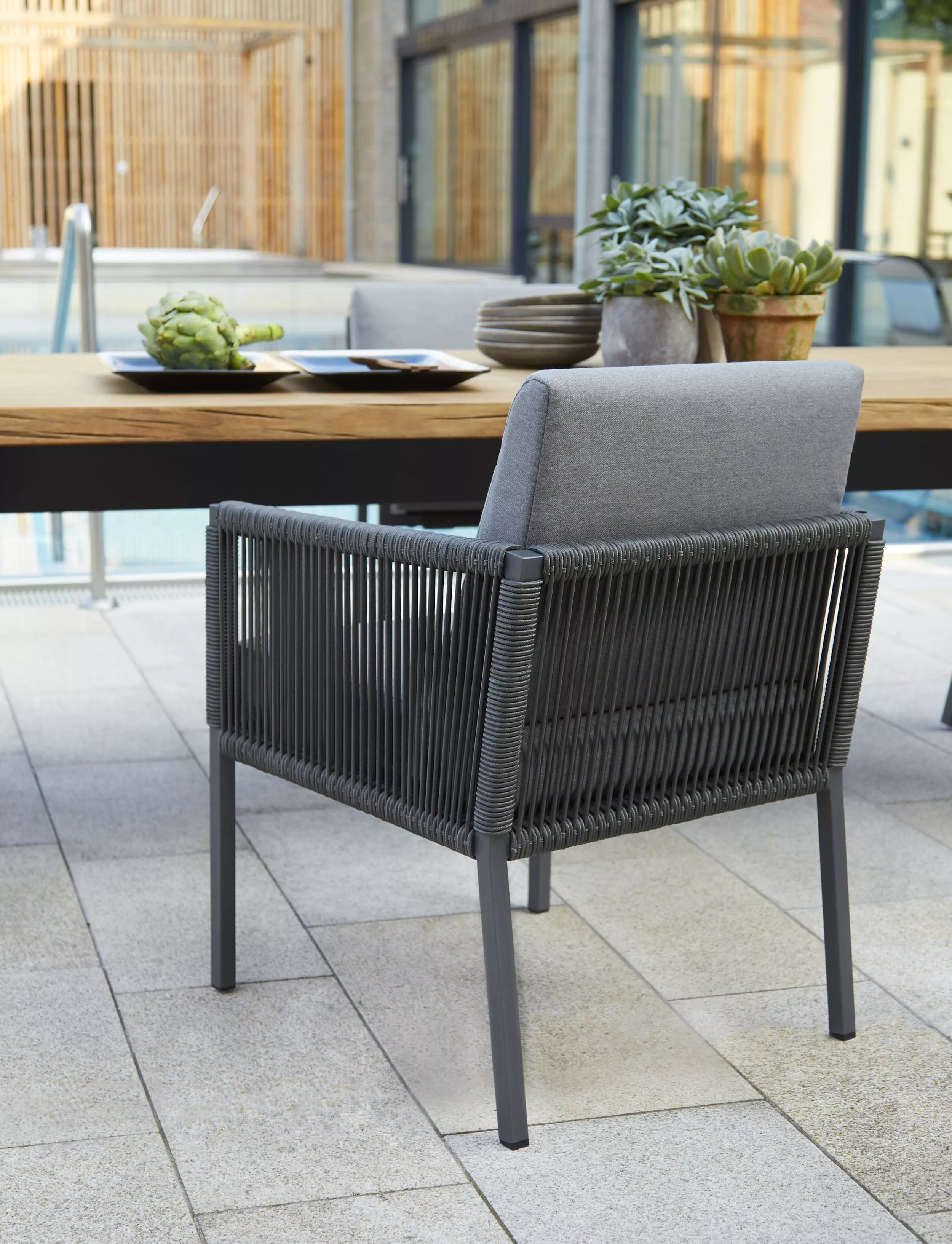 Solpuri Club Ambiente Timber Stuhl mit grauem Polster und schwarzem Gestell steht auf einer Terrasse neben einem Tisch mit Pflanzen und Artischocken.