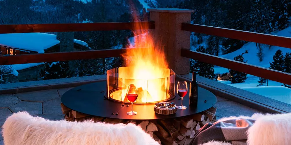 Zwei Weingläser und eine Flasche Wein stehen auf einem Ak-47 Feuerstellen Zero Tisch mit brennendem Feuer im Hintergrund auf einer Terrasse in den Bergen