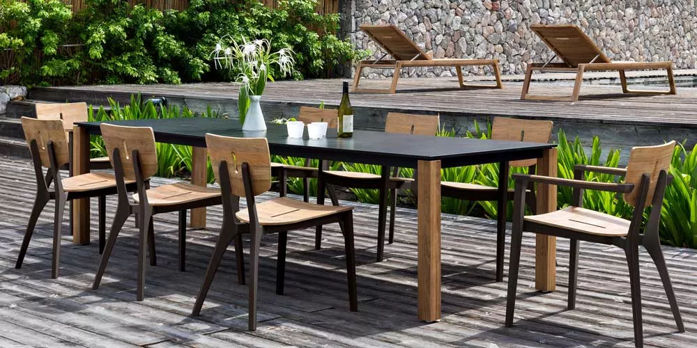 Oasiq Diuna Essgruppe in Naturholz und Schwarz auf einer Terrasse mit Steinmauer und Liegestühlen im Hintergrund.