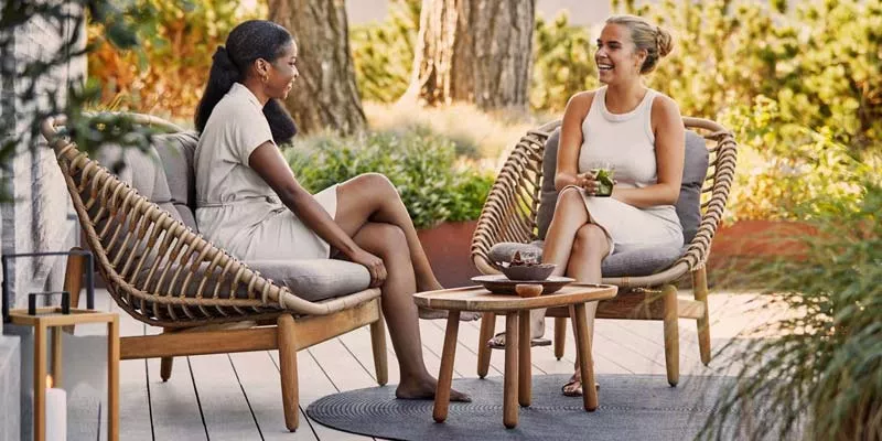 Zwei Frauen sitzen entspannt auf der Cane-line Strington Loungegruppe in natürlichem Holzton auf einer Terrasse im Grünen.