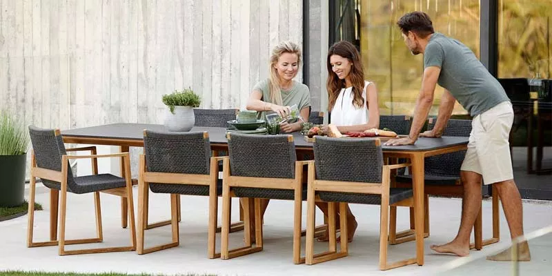 Drei Personen sitzen an einem Cane-line Endless Essgruppe in natürlicher Umgebung, bestehend aus einem Holztisch und passenden Stühlen.