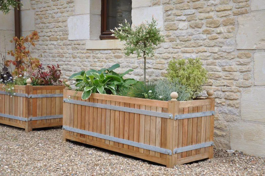Zwei Orangerie Pflanzkübel aus massivem Akazienholz stehen vor einer Steinmauer und sind mit verschiedenen Pflanzen und Sträuchern bepflanzt.