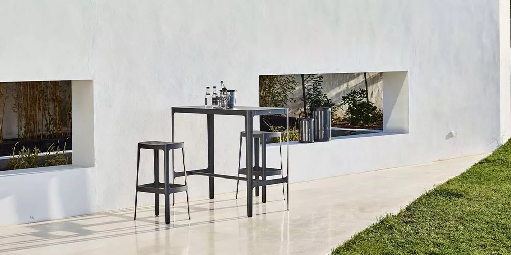 Cane-line Cut Kollektion in Schwarz, bestehend aus einem hohen Tisch und zwei Hockern, auf einer Terrasse vor einer weißen Wand.