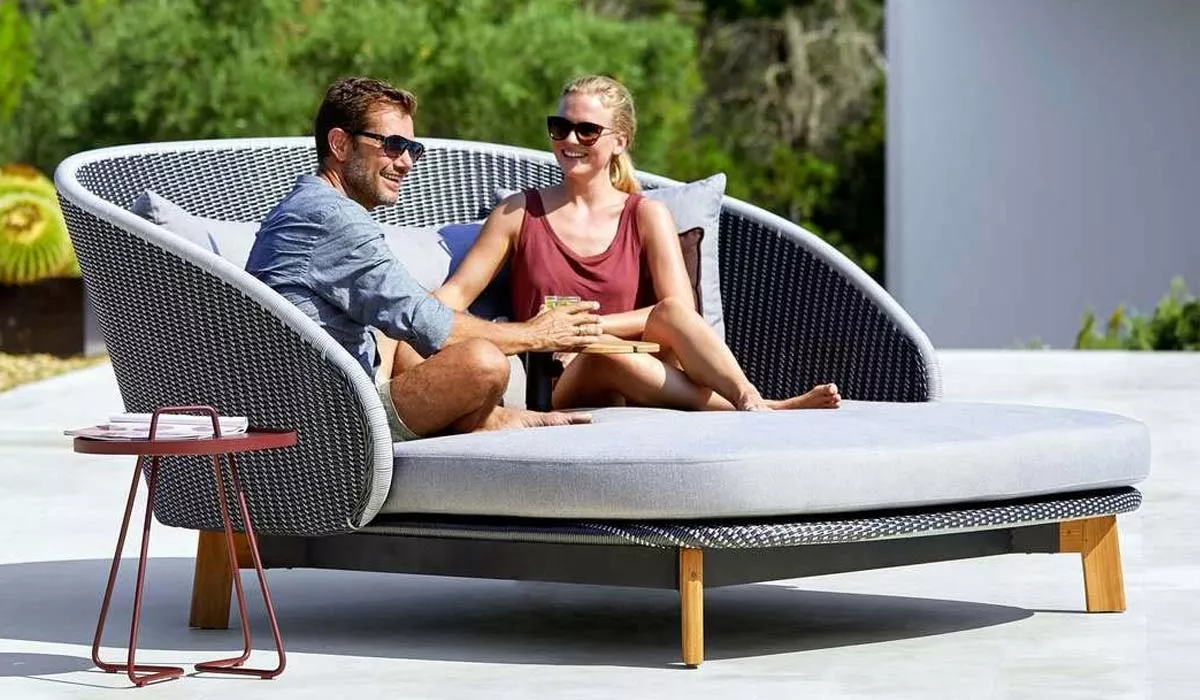 Zwei Personen entspannen auf dem grauen Cane-line Peacock Outdoor Daybed mit Holzbeinen, umgeben von grüner Natur im Hintergrund.