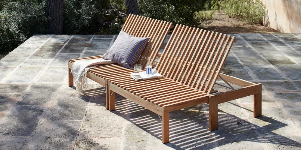 Zwei Skagerak Riviera Sonnenliegen aus Teakholz auf einer Terrasse, mit Kissen und Decke, in einer sonnigen Gartenumgebung.