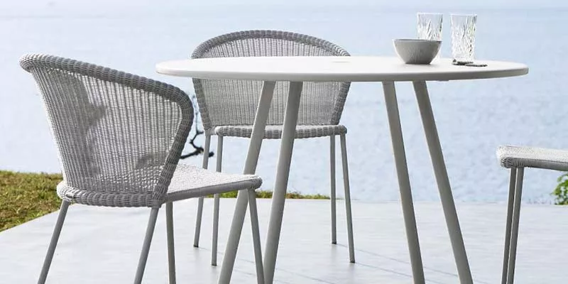 Cane-line Lean Essgruppe in Grau auf einer Terrasse mit Meerblick, bestehend aus einem runden Tisch und zwei Stühlen, in entspannter Atmosphäre.