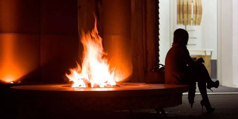 Eine Person sitzt abends neben einer brennenden Ak-47 Feuerstellen Ercole Rain Forest Feuerstelle und genießt die Wärme.