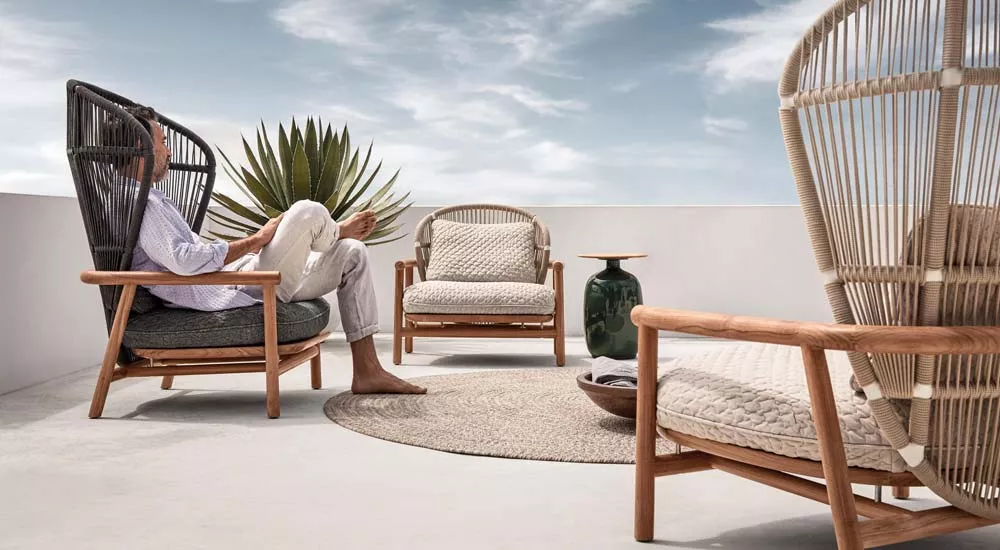 Mann entspannt auf Gloster Fern Gartenmöbel Loungegruppe in Naturtönen auf einer Terrasse mit blauem Himmel im Hintergrund.