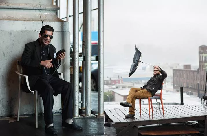 Zwei Männer sitzen auf Dedon Play Essstühlen einer lacht und hält ein Handy der andere kämpft mit einem Regenschirm im Freien auf einer Terrasse.