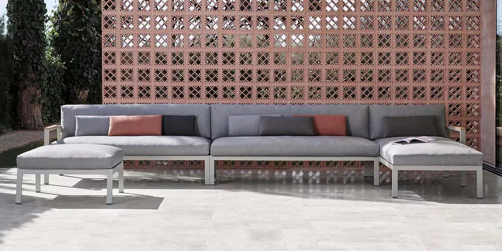 Gandia Blasco Timeless Sofa in Grau auf einer Terrasse vor einer gemusterten Ziegelwand, mit Kissen in verschiedenen Farben.