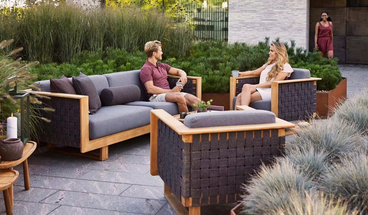 Zwei Personen entspannen auf einer Cane-line Loungegruppe in Grau und Holzoptik im Garten, umgeben von grüner Vegetation.