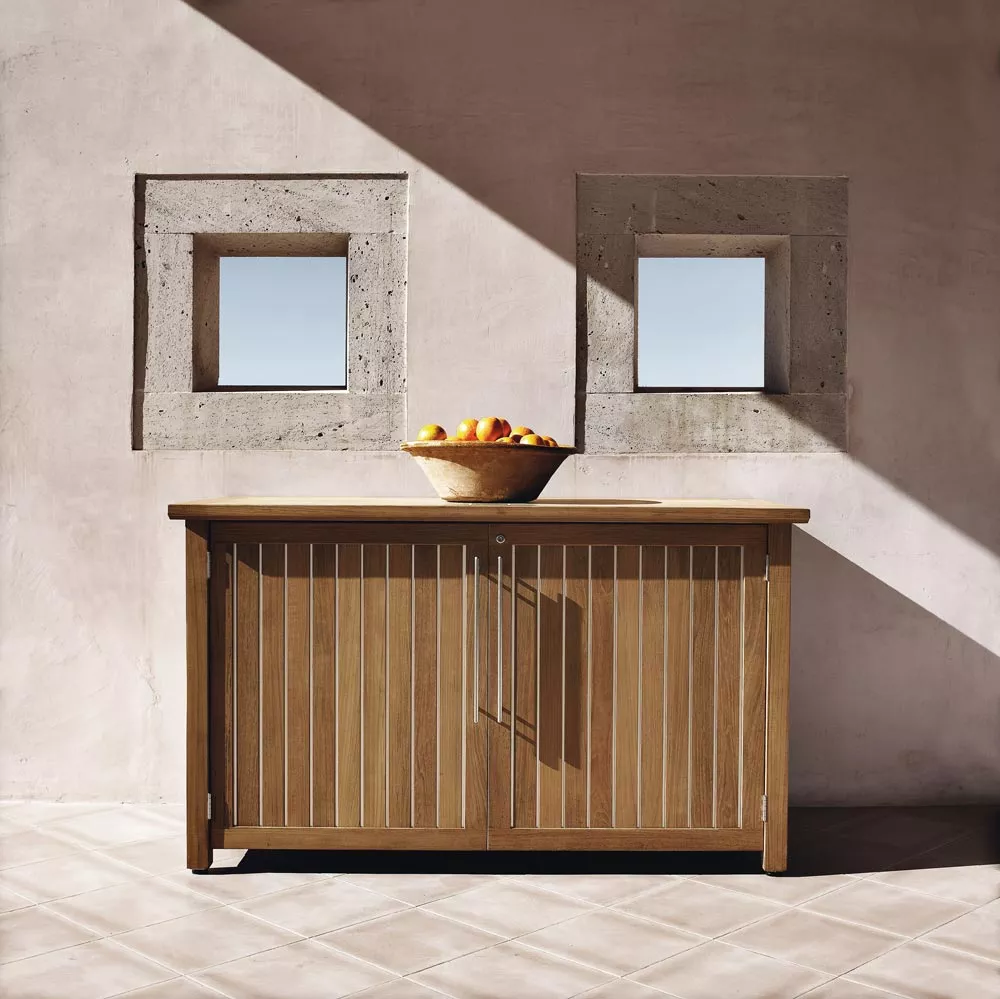 Kissentruhe aus Teakholz von Gloster, naturfarben, vor einer Wand mit zwei Fenstern, im Sonnenlicht.