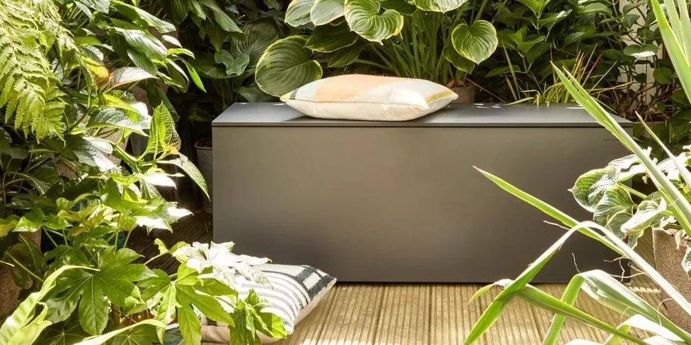 Conmoto Kollektion ELPECHO, anthrazit, Outdoor-Sitzbank in einem üppigen Garten, umgeben von grünen Pflanzen, mit Kissen dekoriert.