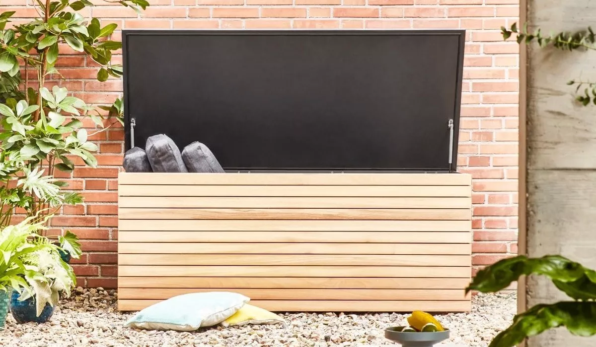 Conmoto Kissentruhe in hellem Holz vor einer Ziegelwand, geöffnet mit grauen Kissen, umgeben von Pflanzen auf Kiesboden.