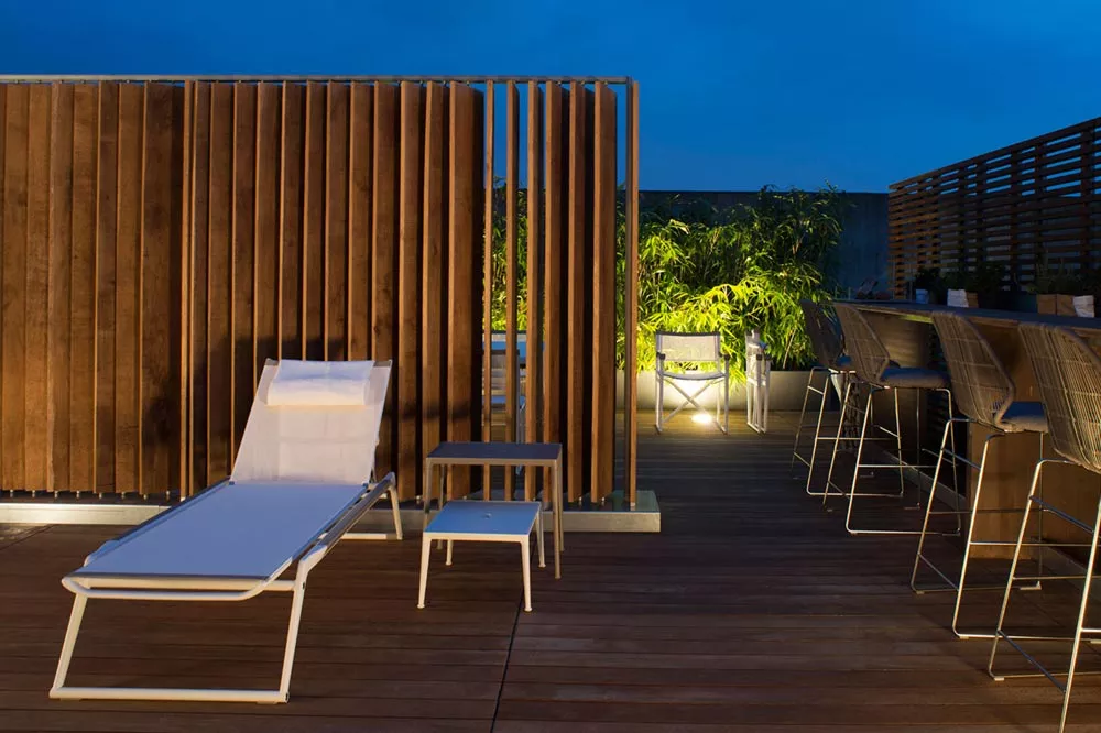 B&B Italia Mirto Outdoor Sonnenliege in weiß auf einer stilvollen Terrasse bei Nacht mit Holzwänden und Pflanzen im Hintergrund