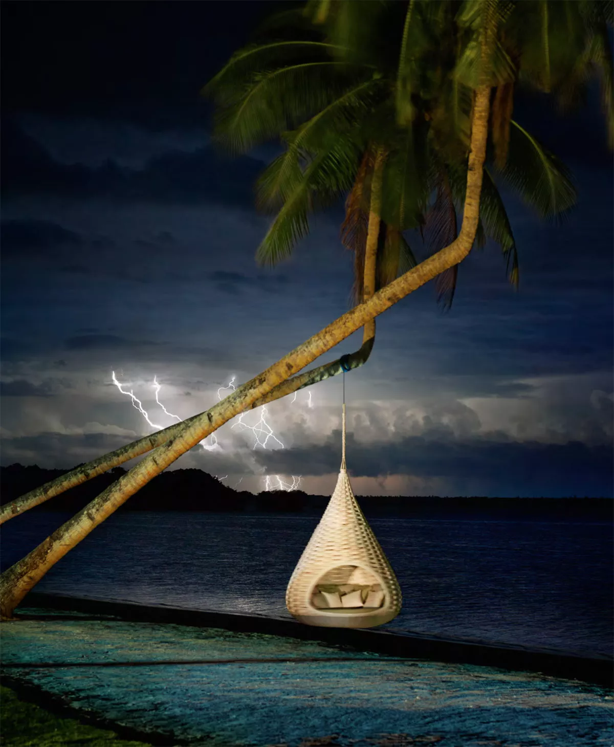 Nestrest Hanging Lounger rund 200 cm von Dedon hängt an einem Baum am Strand während eines Gewitters im Hintergrund blitzen Blitze.