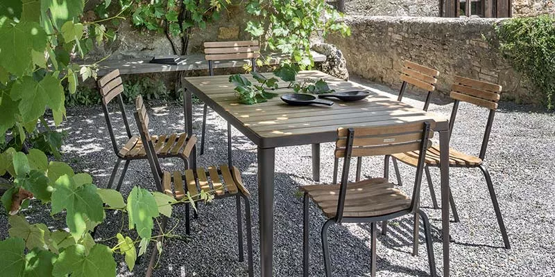 Ethimo Laren Outdoor Ess-Loungegruppe in Holzoptik auf Kiesboden, umgeben von grünen Pflanzen und Steinmauer im Hintergrund.