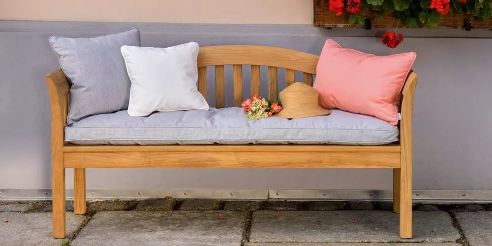 Weißhäupl Victorian Bank aus Holz mit grauem Sitzkissen, dekoriert mit bunten Kissen und Strohhut, vor einer Hauswand.