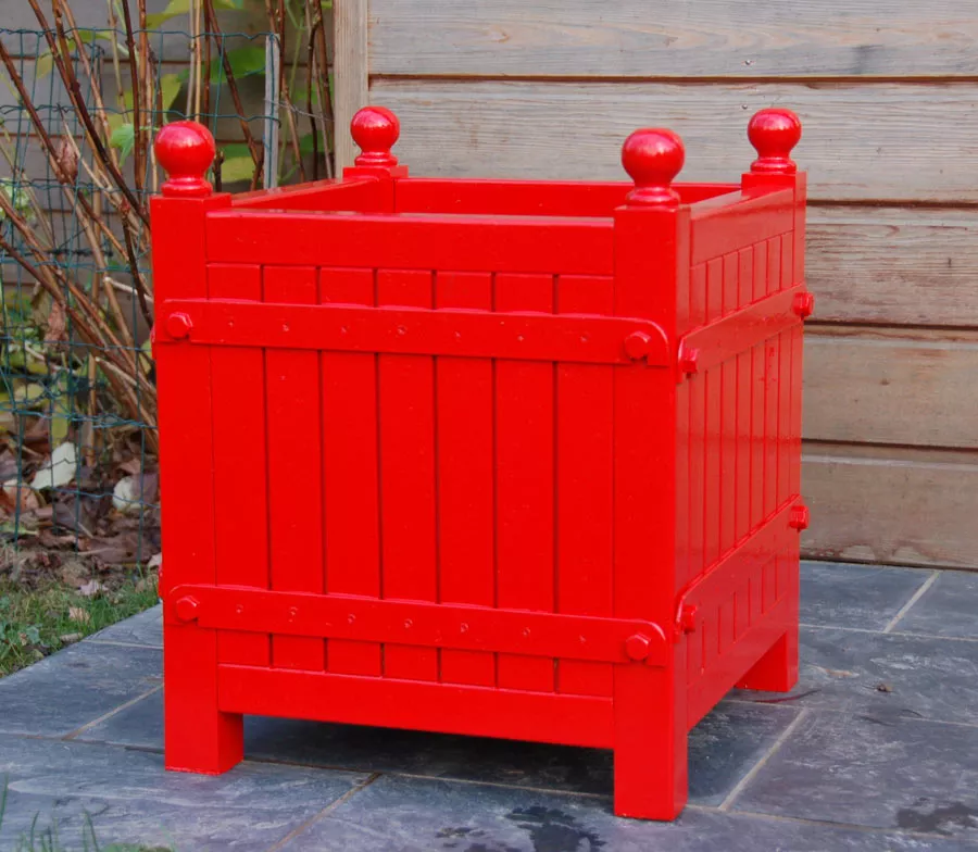 Orangerie Pflanzkübel aus lackiertem Akazienholz in leuchtendem Rot auf einer Terrasse vor einer Holzwand im Garten.