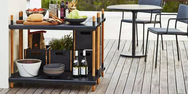 Cane-line Frame Regalsystem in Schwarz und Holzoptik auf einer Terrasse mit gedecktem Tisch im Hintergrund.