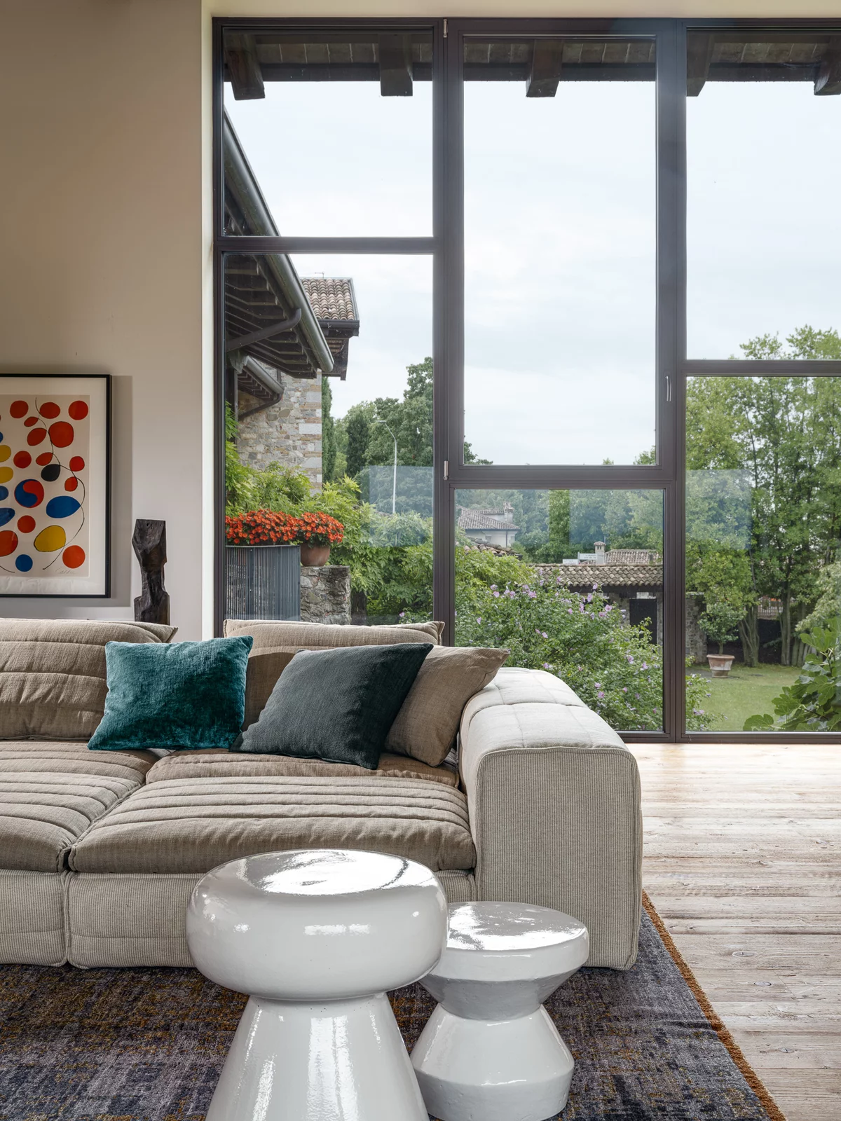 Ein gemütliches Wohnzimmer mit einem beigen Sofa von Gervasoni und zwei weißen Beistelltischen vor einer großen Fensterfront mit Blick ins Grüne.