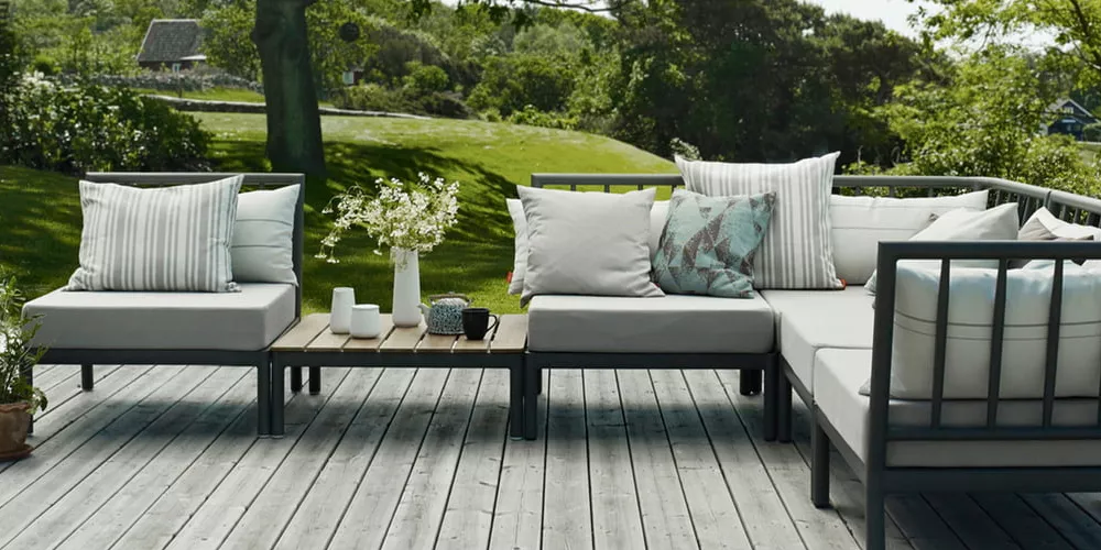 Gartenmöbel-Set von Skagerak in Grau auf einer Holzterrasse mit grüner Wiese im Hintergrund, dekoriert mit Kissen und Blumen.
