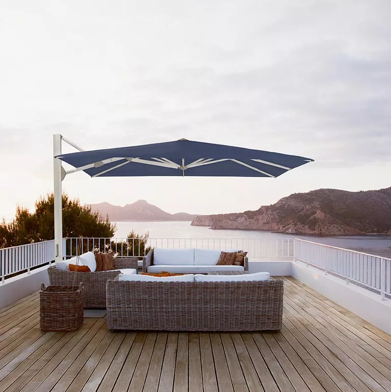 Gastronomie-Sonnenschirm Ambiente Nova von Glatz in Blau auf einer Terrasse mit Meerblick und gemütlichen Rattanmöbeln bei Sonnenuntergang.