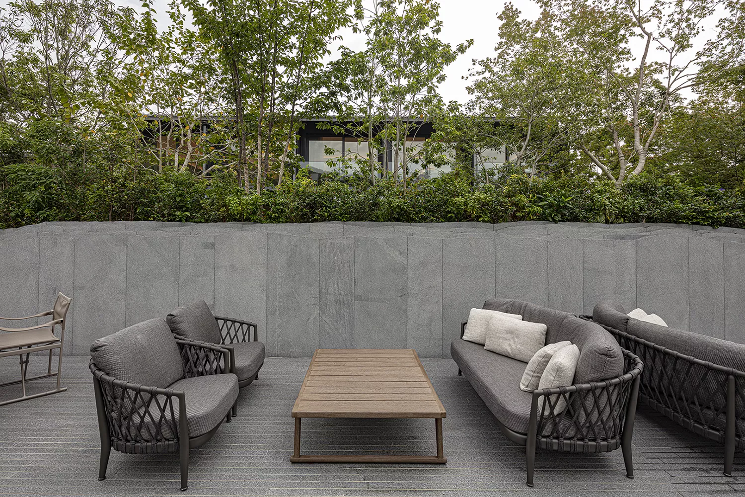 B&B Italia Erica Loungesofa 216 in einem modernen Garten mit grauen Kissen und einem Holztisch in der Mitte vor einer Steinmauer und Bäumen im Hintergrund