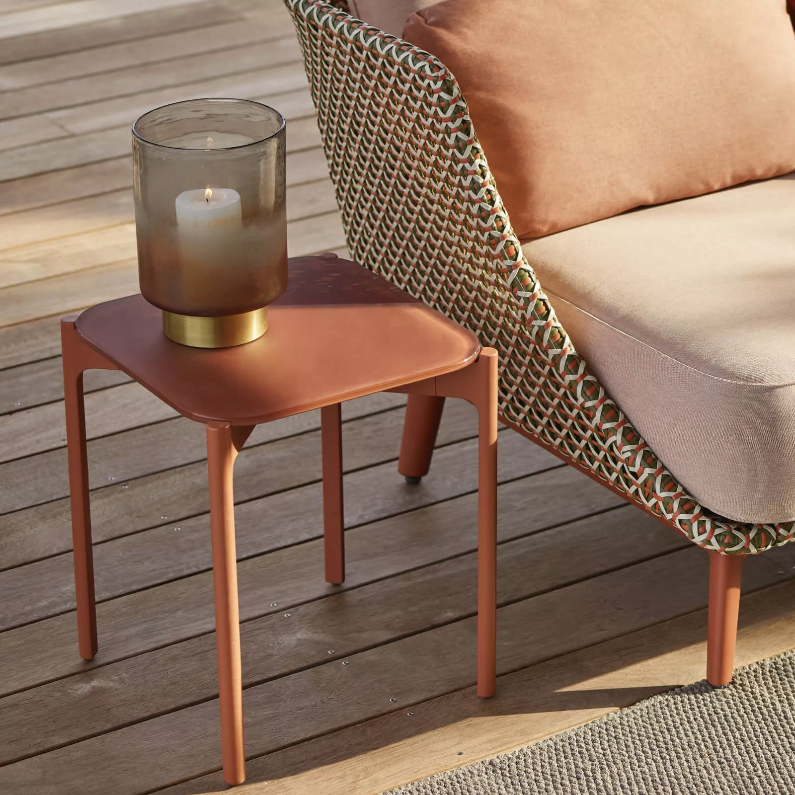 Dedon Izon Kollektion Beistelltisch in Terrakotta mit Kerze auf einer Terrasse neben einem geflochtenen Sessel mit Kissen