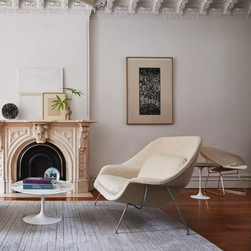 Saarinen Womb Settee in Beige in einem eleganten Wohnzimmer mit Kamin und Kunstwerken an der Wand, auf einem grauen Teppich.
