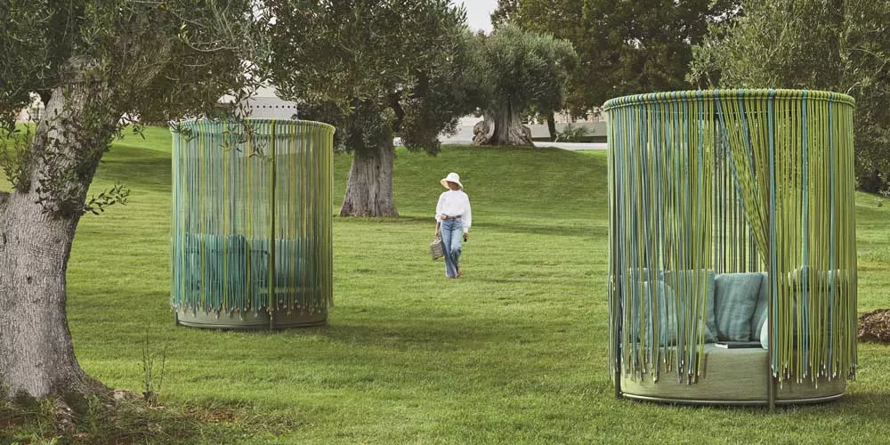 Zwei grüne Roberti Oasi Gartenmöbel auf einer Wiese, umgeben von Bäumen, mit einer Person im Hintergrund, die einen Hut trägt.