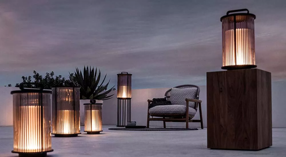 Gloster Ambient Gartenleuchten in warmem Licht auf einer Terrasse bei Dämmerung, umgeben von Pflanzen und einem Holzstuhl.