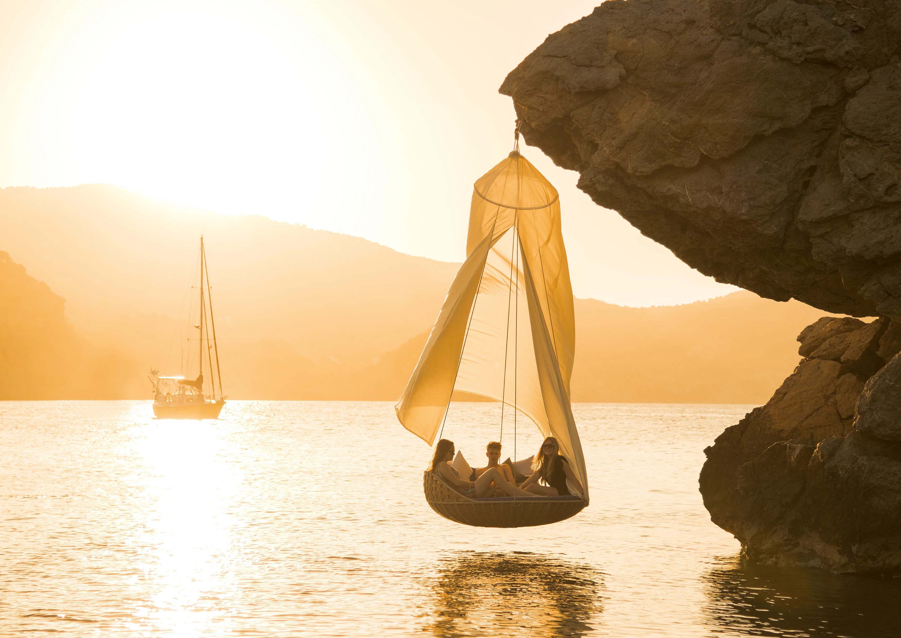 Drei Personen entspannen in einem Dedon Swingrest Hängesessel der an einer Klippe hängt während die Sonne über dem Meer untergeht und ein Boot vorbeifährt
