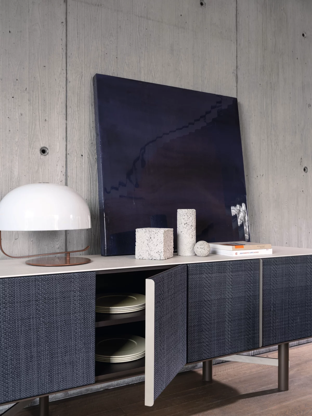 Gervasoni Sideboard mit blauen Türen und weißer Tischlampe auf einer Holzplatte vor einer Betonwand mit Kunstwerk und Dekoration