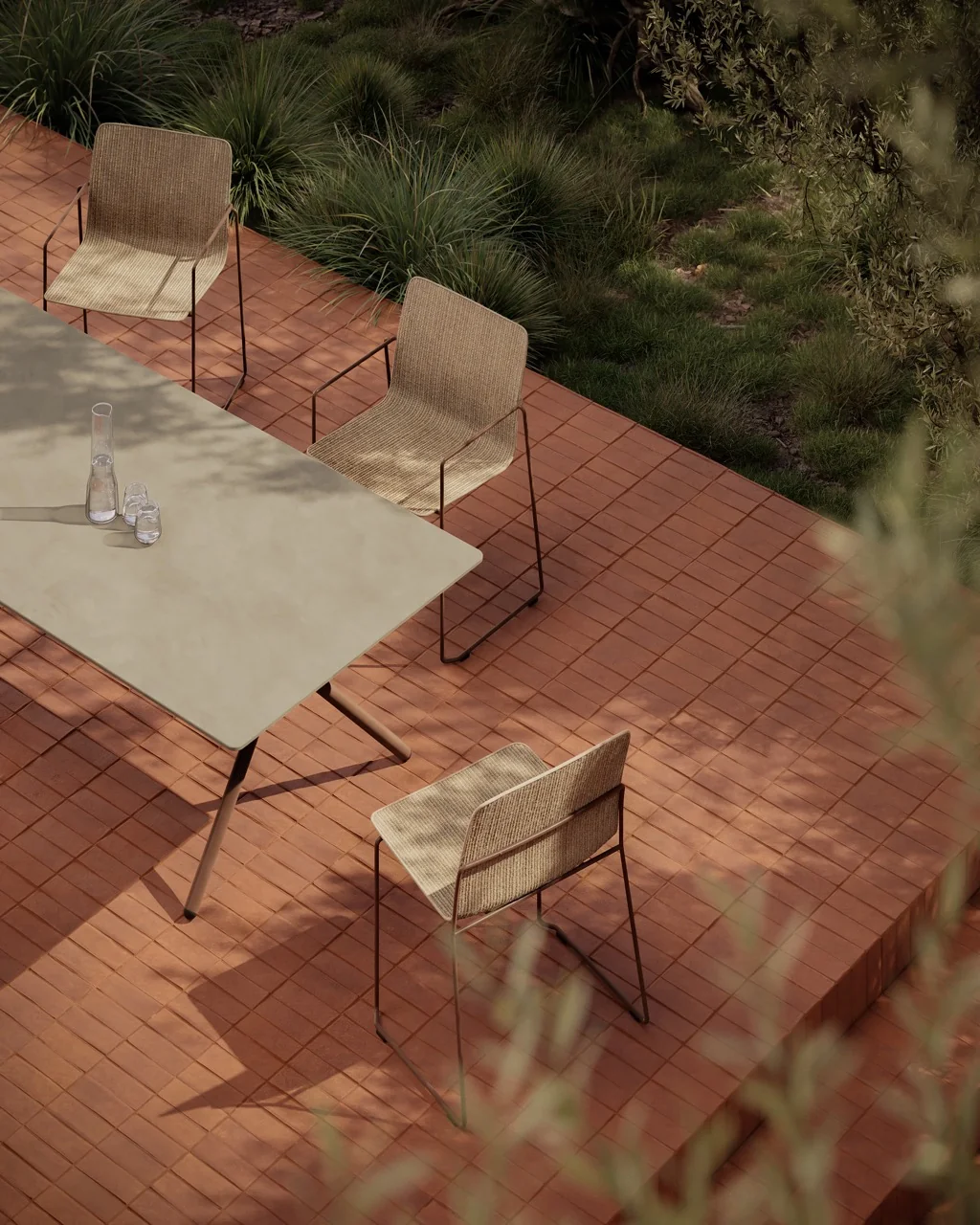 Bivaq Sal Kollektion: Vier beige Stühle und ein Tisch auf einer roten Terrassenfliese, umgeben von grüner Vegetation, stimmungsvolles Bild.