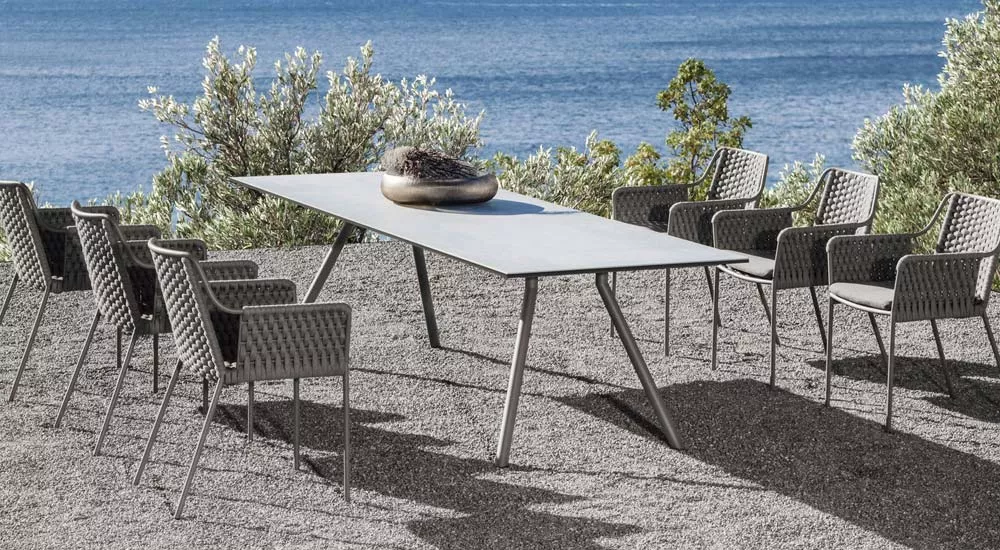 Fischer Möbel Wing Teso Essgruppe in Grau auf Kiesboden mit Meerblick, bestehend aus Tisch und acht Stühlen, Outdoor-Setting.