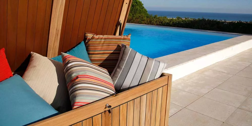 Barlow Tyrie Aufbewahrungstruhe aus Teakholz mit bunten Kissen, im Hintergrund ein Pool mit Meerblick bei sonnigem Wetter.
