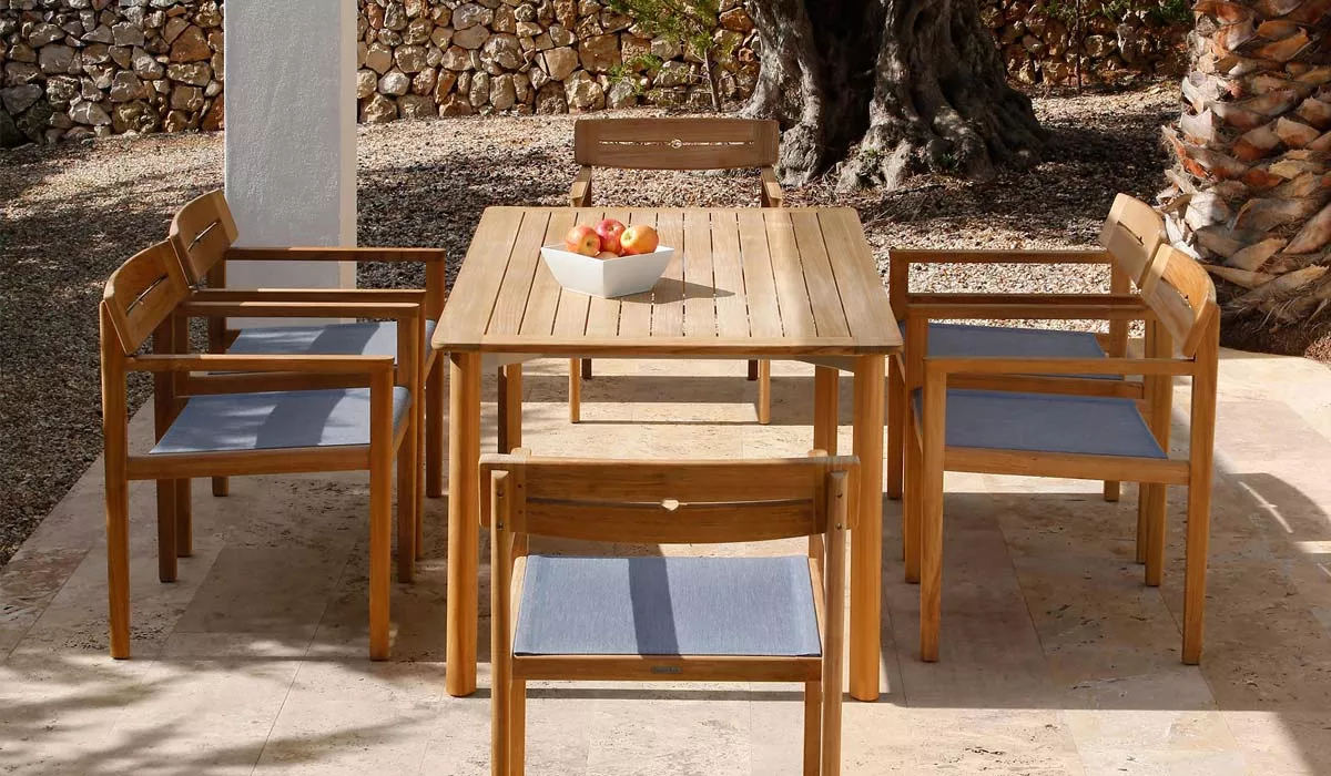 Barlow Tyrie Atom Essgruppe aus Teakholz mit blauen Sitzpolstern auf einer Terrasse vor einer Steinmauer und einem Baum.