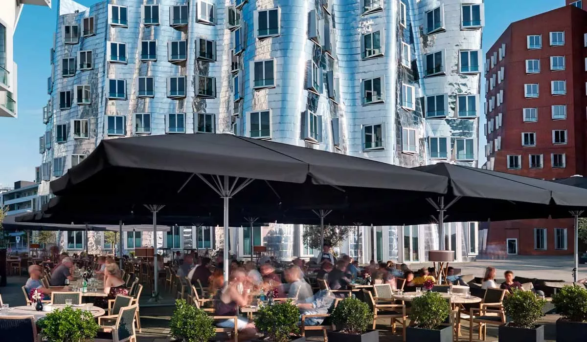 Schwarzer May Schattello Sonnenschirm auf einer belebten Terrasse vor moderner Architektur, stimmungsvolle Außenaufnahme.